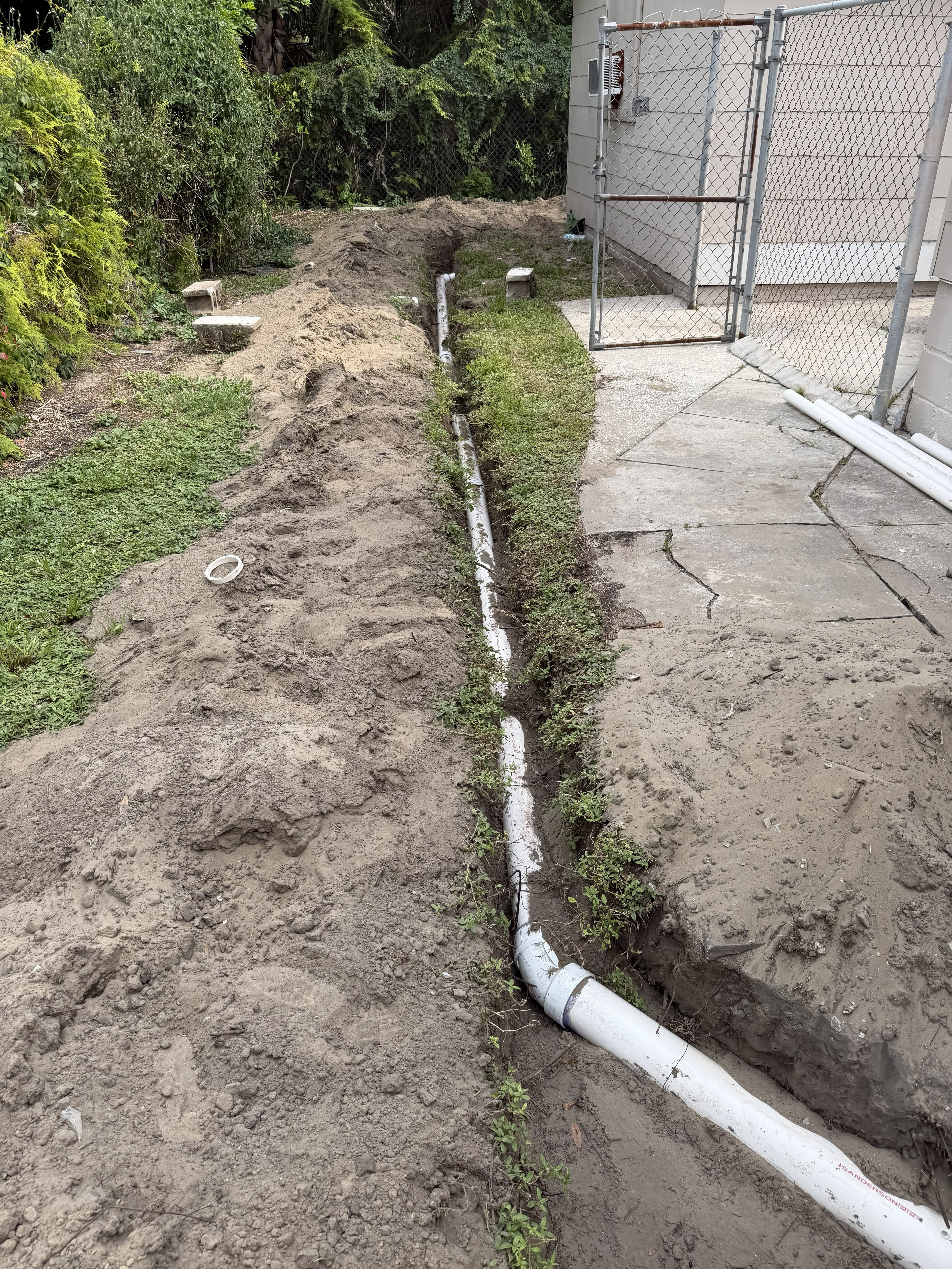 New white PVC sewer lateral with two-way cleanout installed in an open trench, old rusted pipe sections piled beside it